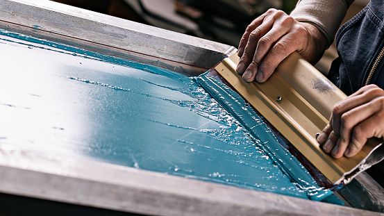 Rodo espalhando tinta azul sobre a tela de silk screen