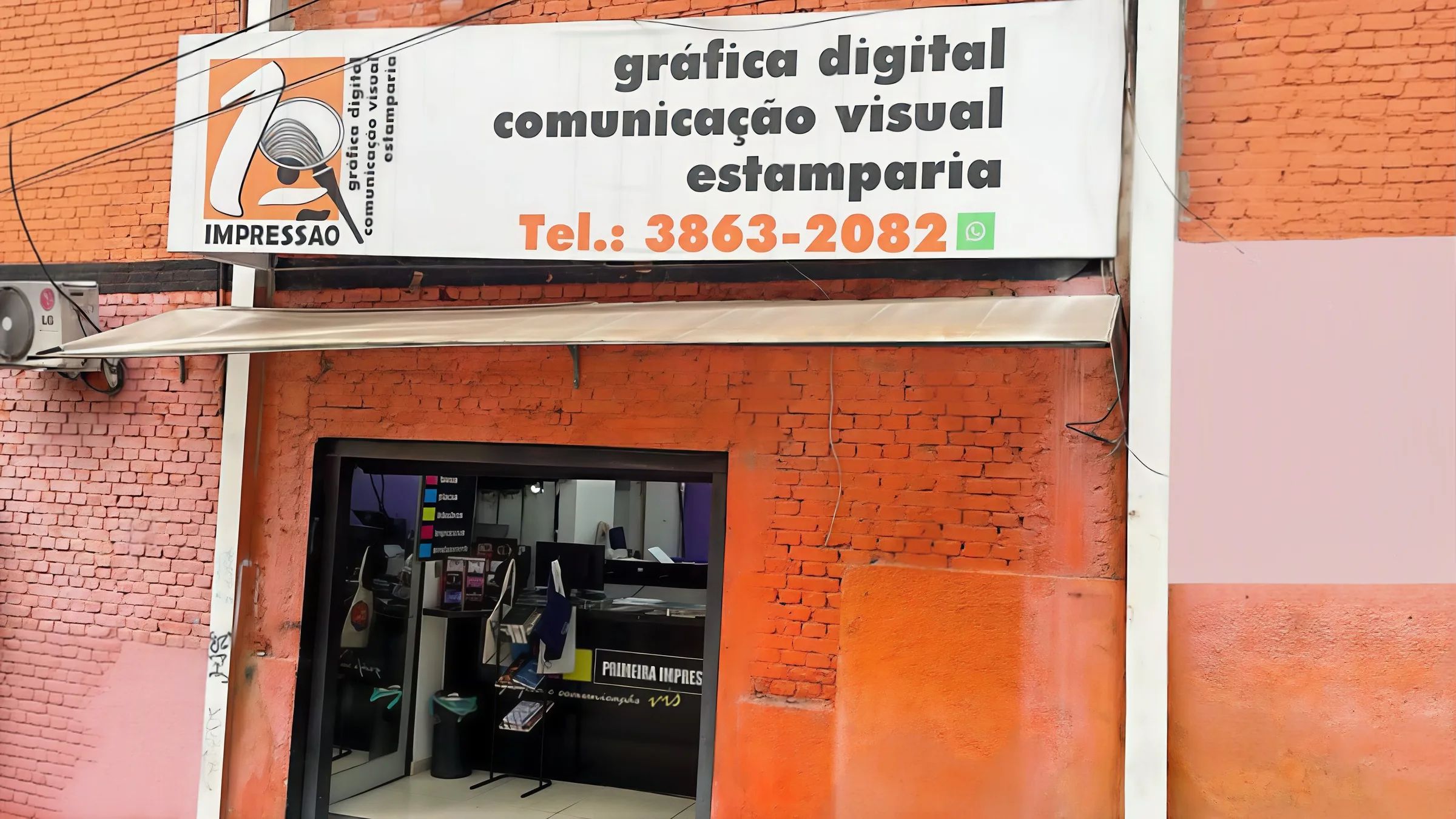 Fachada da Primeira Impressão: parede de tijolos laranja, placa branca com 'gráfica digital, comunicação visual, estamparia' e o telefone, porta de vidro aberta mostrando o balcão.